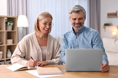 Photo of Happy middle aged couple working with laptop at wooden table indoors