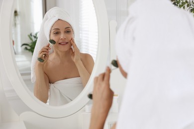 Photo of Smiling woman doing facial self massage with roller near mirror in bathroom