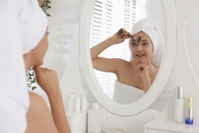 Photo of Smiling woman doing facial self massage with roller near mirror in bathroom