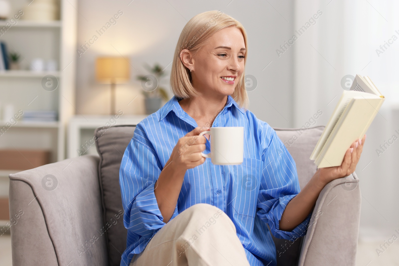 Photo of Smiling middle aged woman with cup of hot drink reading book at home