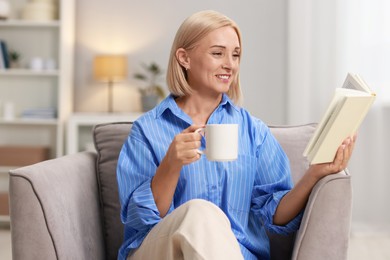 Smiling middle aged woman with cup of hot drink reading book at home