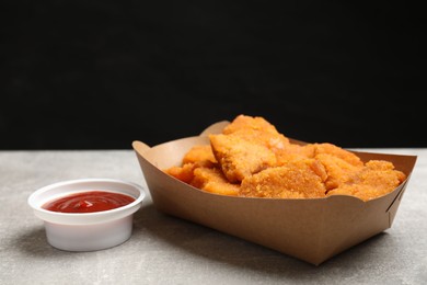 Photo of Delicious chicken nuggets in carton box and ketchup on gray textured table against black background