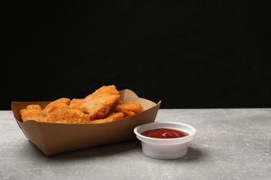 Photo of Delicious chicken nuggets in carton box and ketchup on gray textured table against black background. Space for text