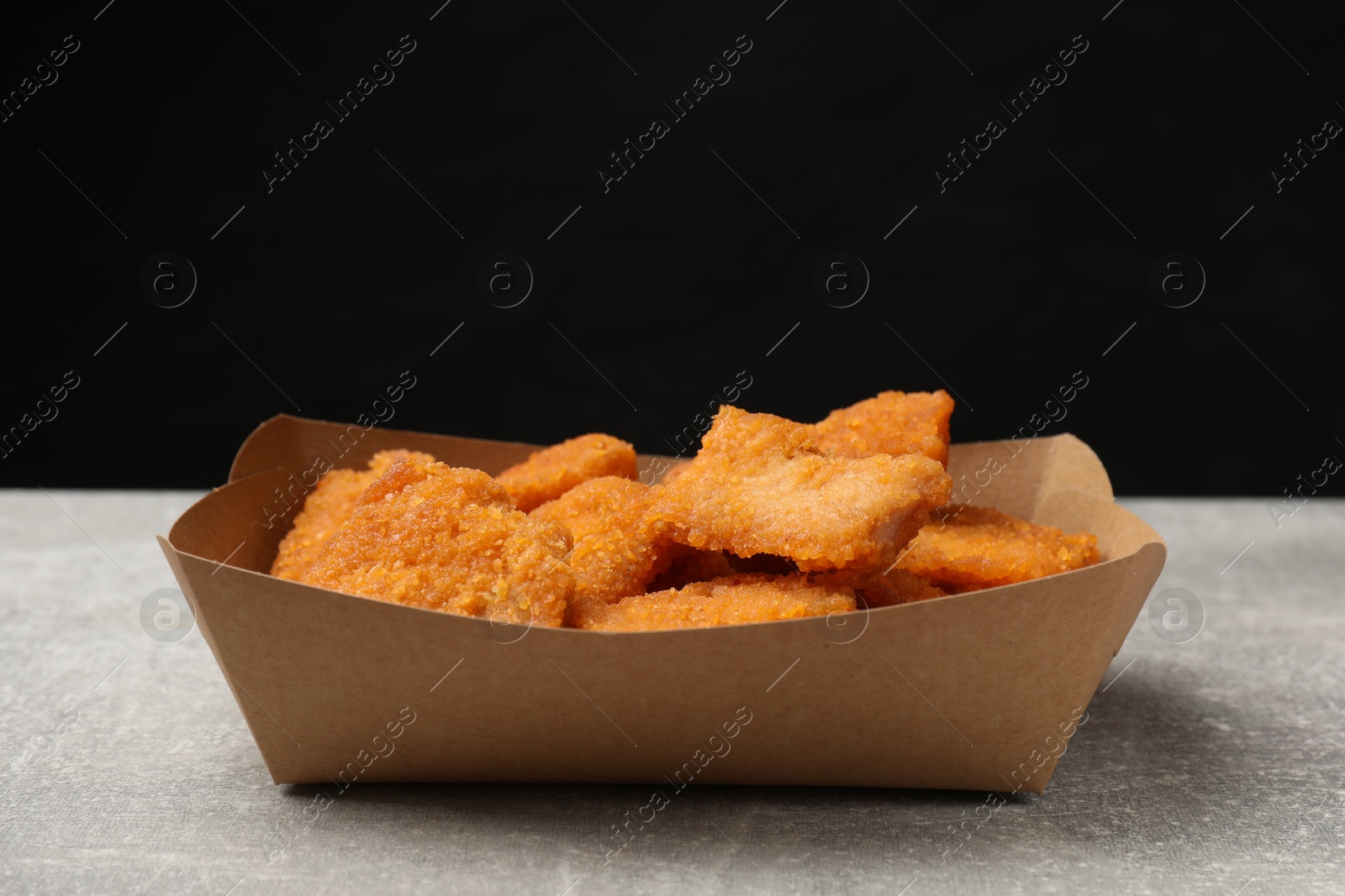 Photo of Delicious chicken nuggets in carton box on gray textured table against black background