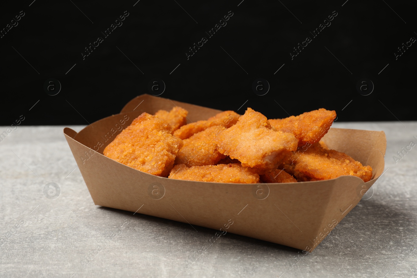 Photo of Delicious chicken nuggets in carton box on gray textured table against black background