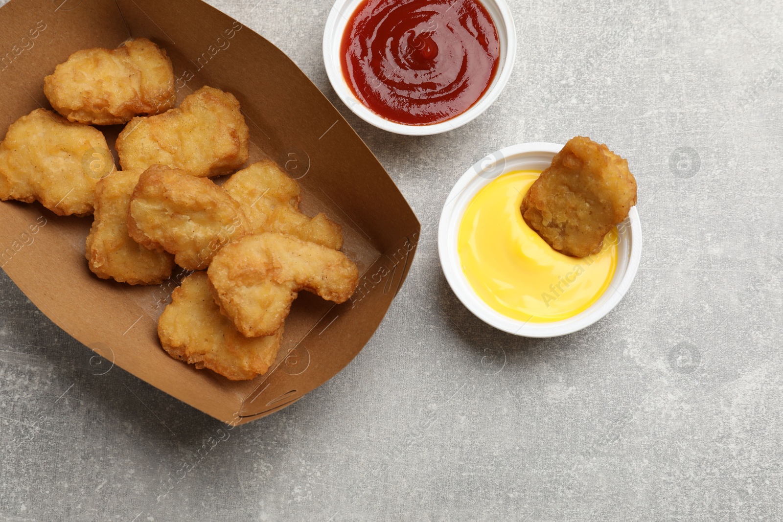 Photo of Delicious chicken nuggets in carton box and sauces on gray textured table, flat lay