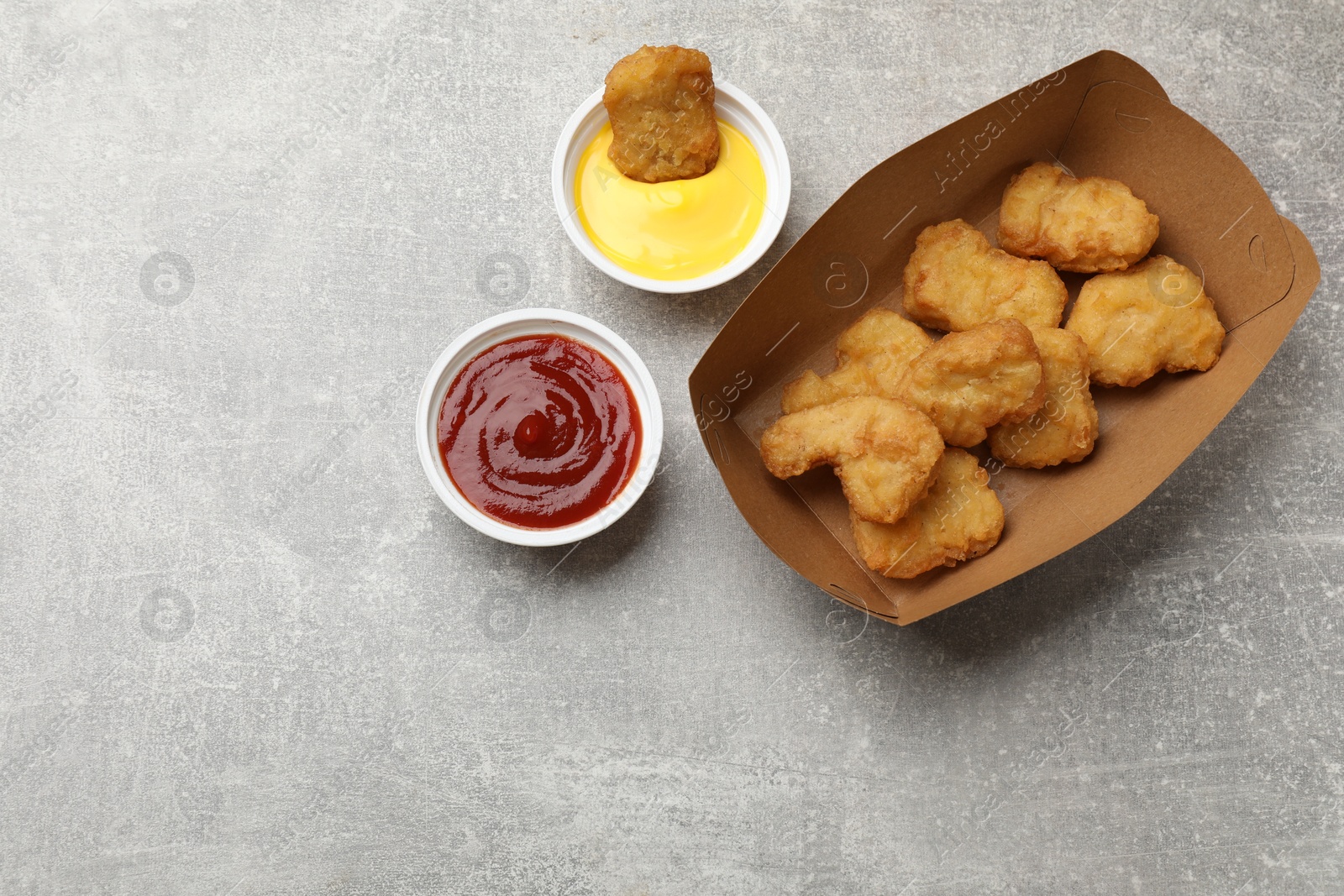 Photo of Delicious chicken nuggets in carton box and sauces on gray textured table, flat lay. Space for text