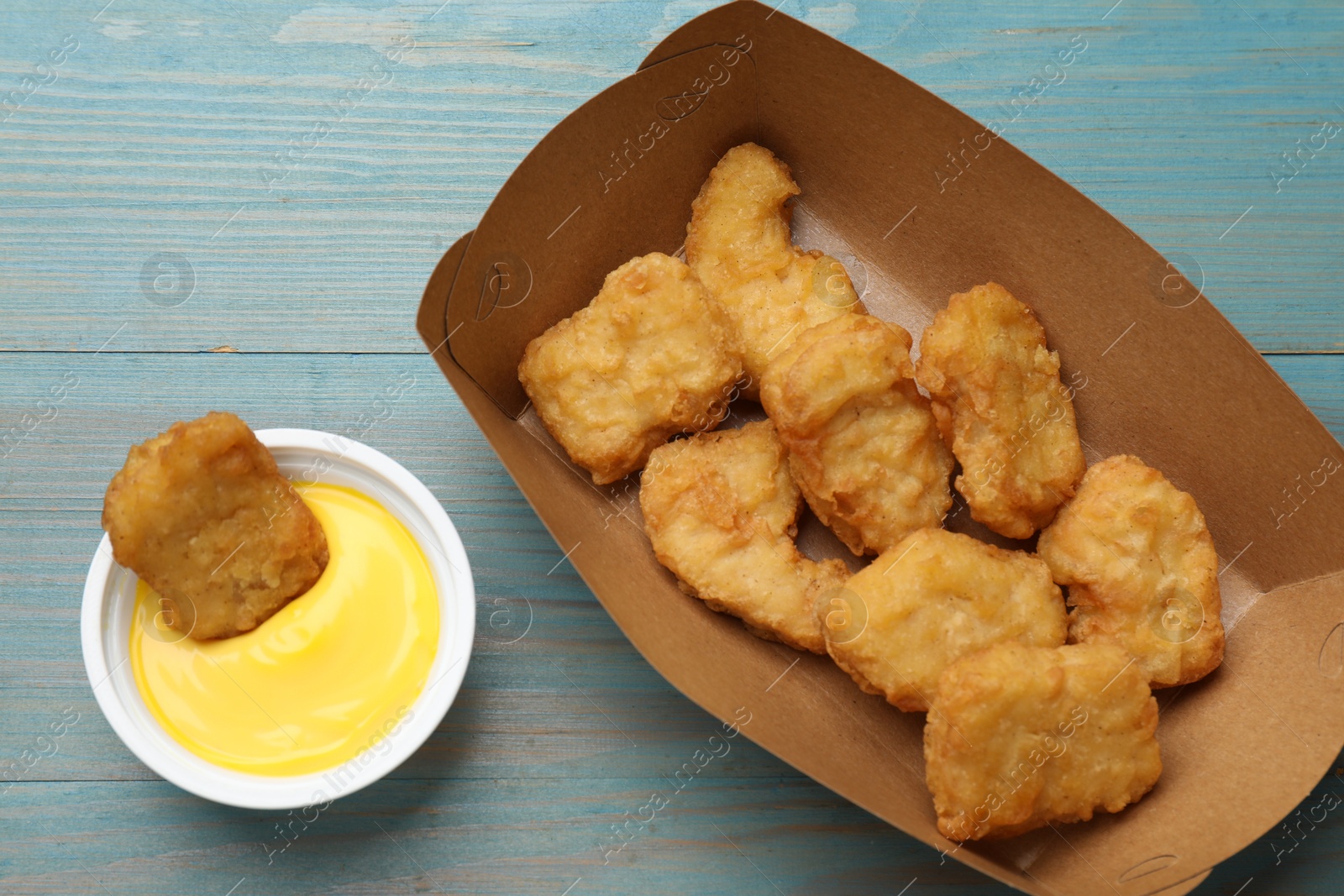 Photo of Delicious chicken nuggets in carton box and sauce on light blue wooden table, flat lay