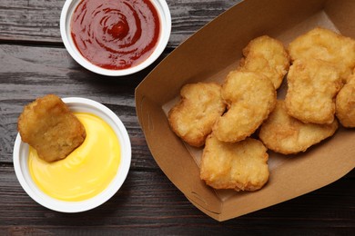 Delicious chicken nuggets in carton box and sauces on wooden table, flat lay