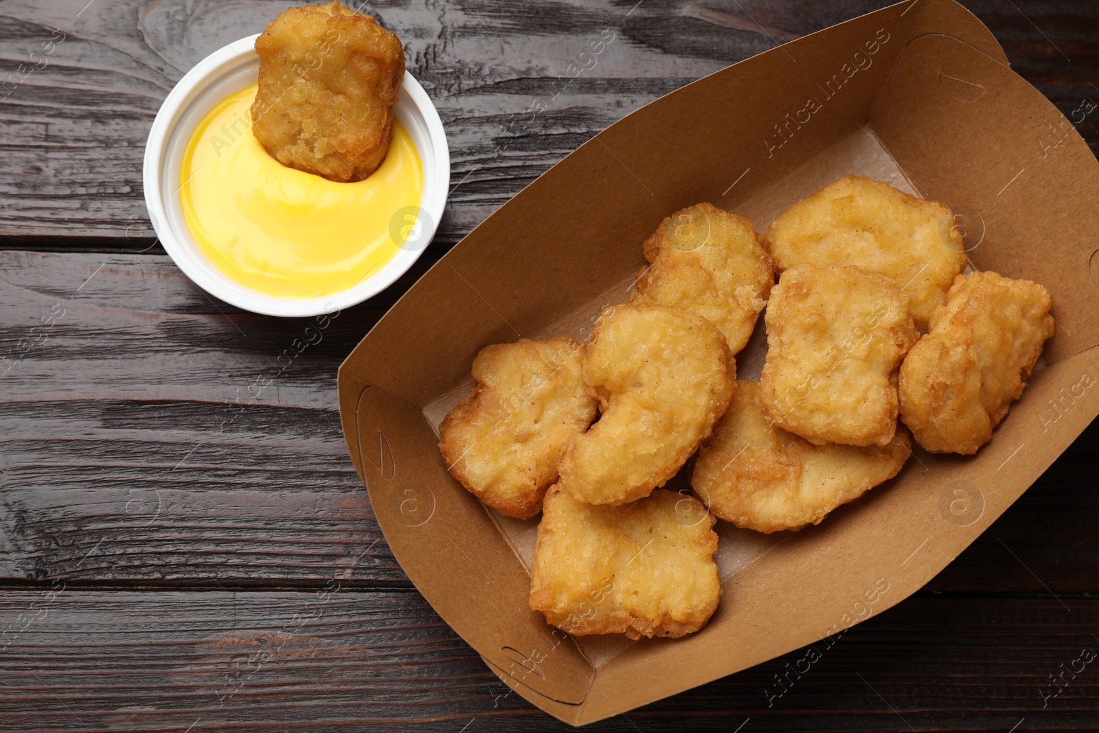 Photo of Delicious chicken nuggets in carton box and sauce on wooden table, flat lay