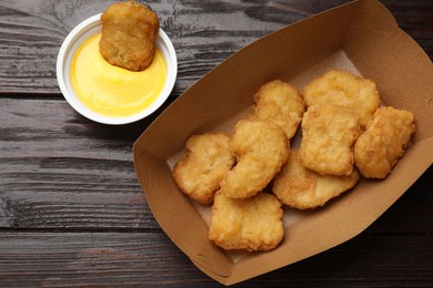 Delicious chicken nuggets in carton box and sauce on wooden table, flat lay