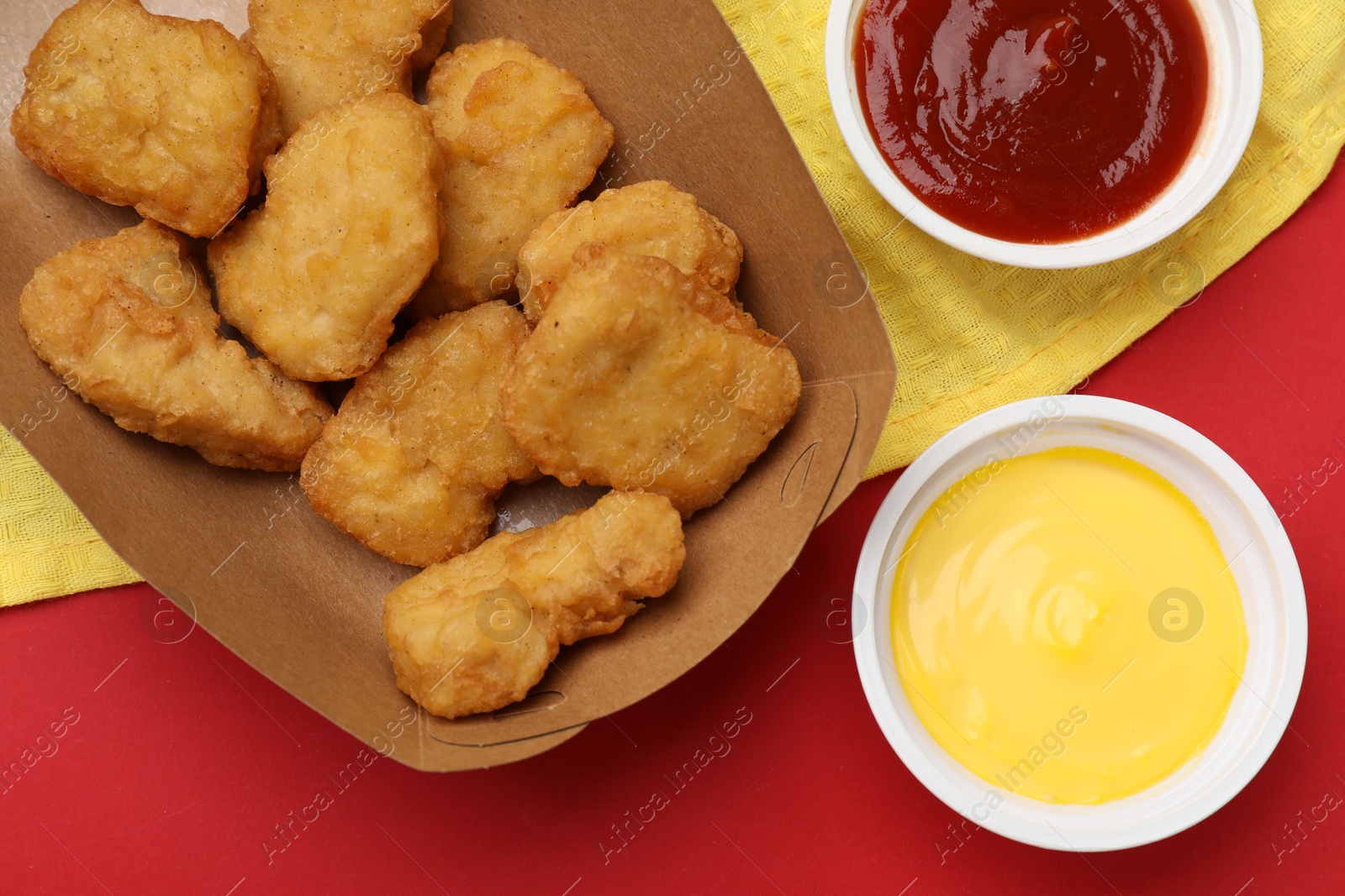 Photo of Delicious chicken nuggets in carton box and sauces on red background, flat lay