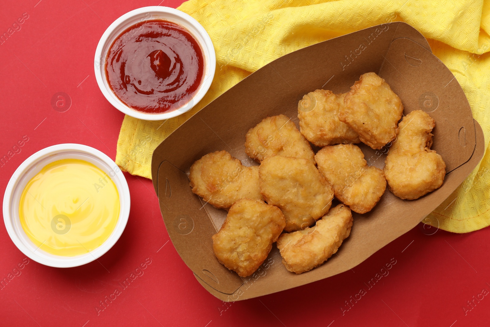 Photo of Delicious chicken nuggets in carton box and sauces on red background, flat lay