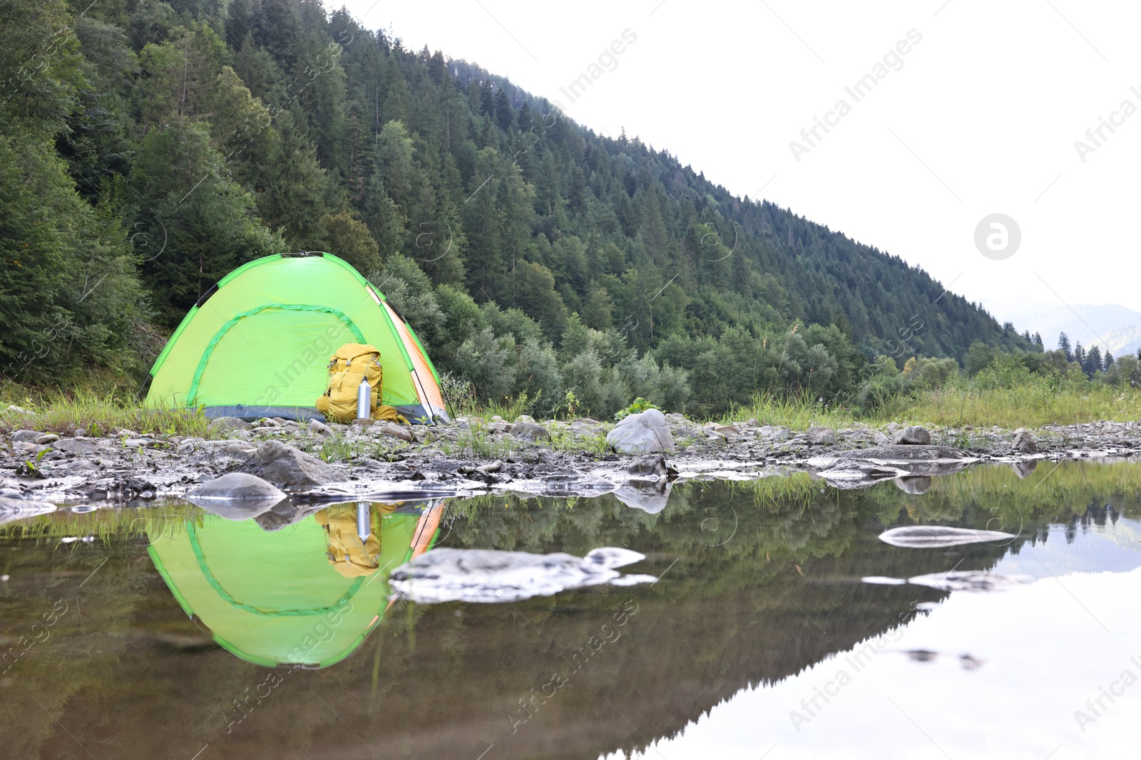 Photo of Camping tent, backpack and thermo bottle on stones near river in mountains