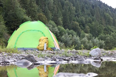 Photo of Camping tent, backpack and thermo bottle on stones near river in mountains