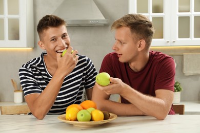 Happy brothers eating apples at table in kitchen