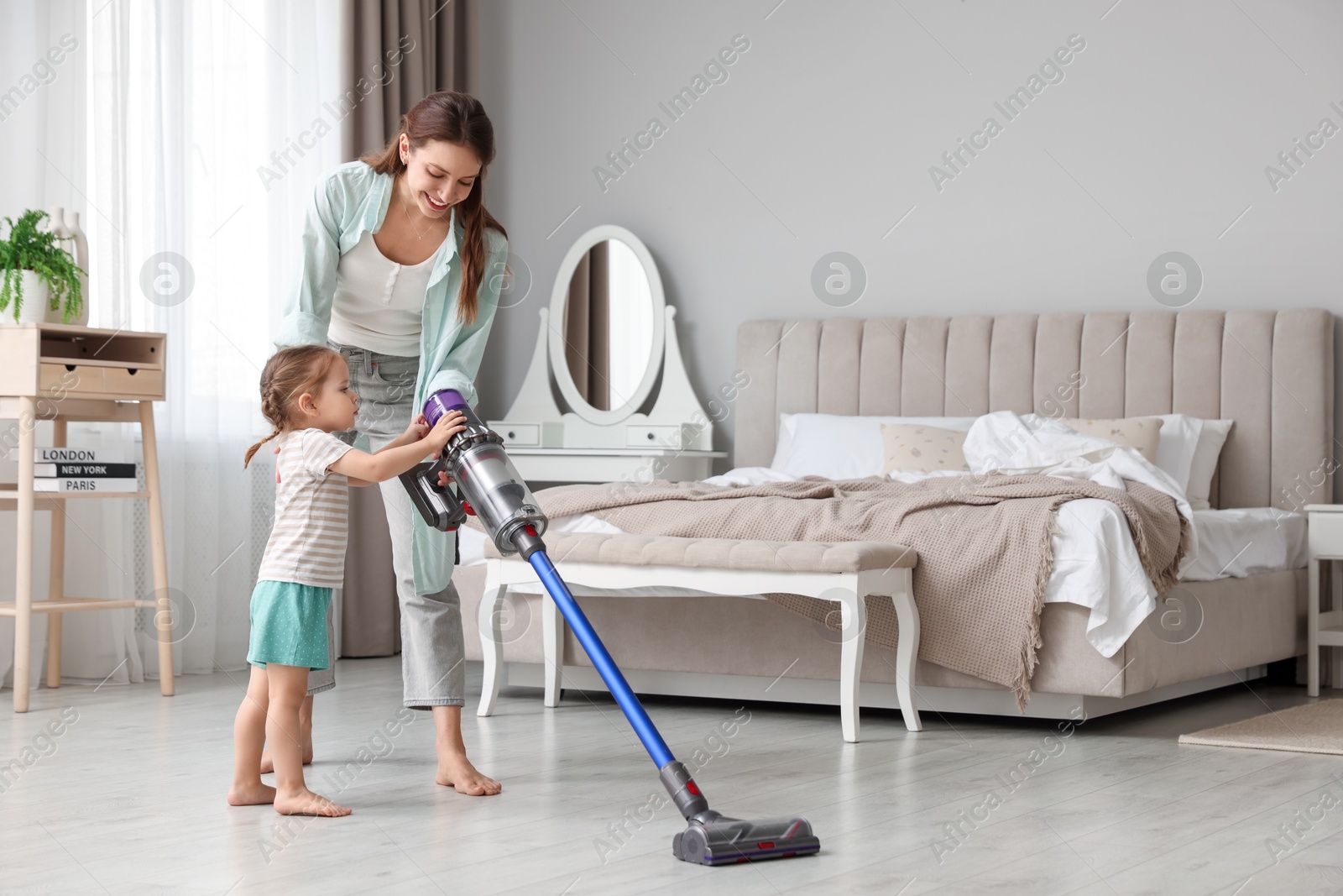 Photo of Smiling young woman and her daughter cleaning floor with cordless vacuum cleaner in bedroom