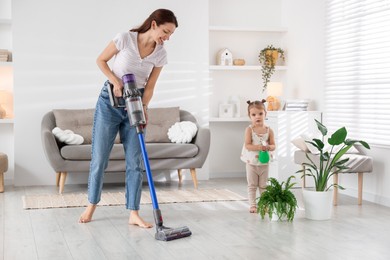 Photo of Smiling young woman cleaning floor with cordless vacuum cleaner while her daughter spraying houseplant at home