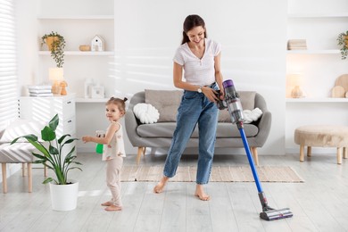 Smiling young woman cleaning floor with cordless vacuum cleaner while her daughter spraying houseplant at home