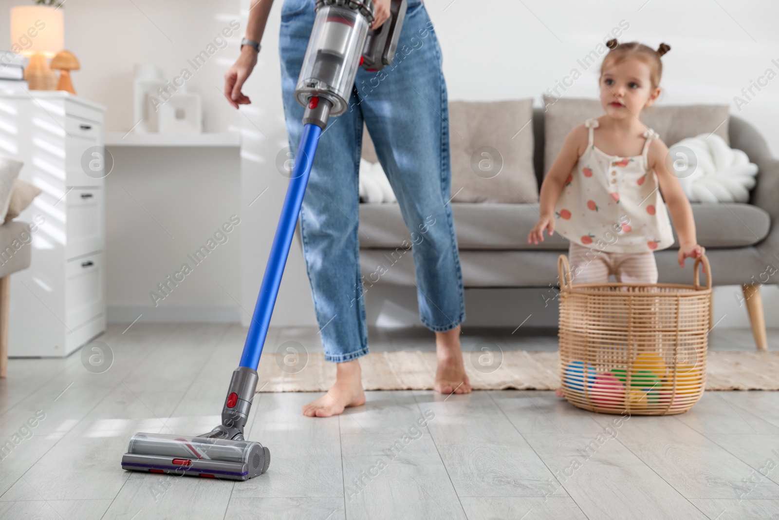 Photo of Young woman cleaning floor with cordless vacuum cleaner while her daughter playing with toys at home, closeup