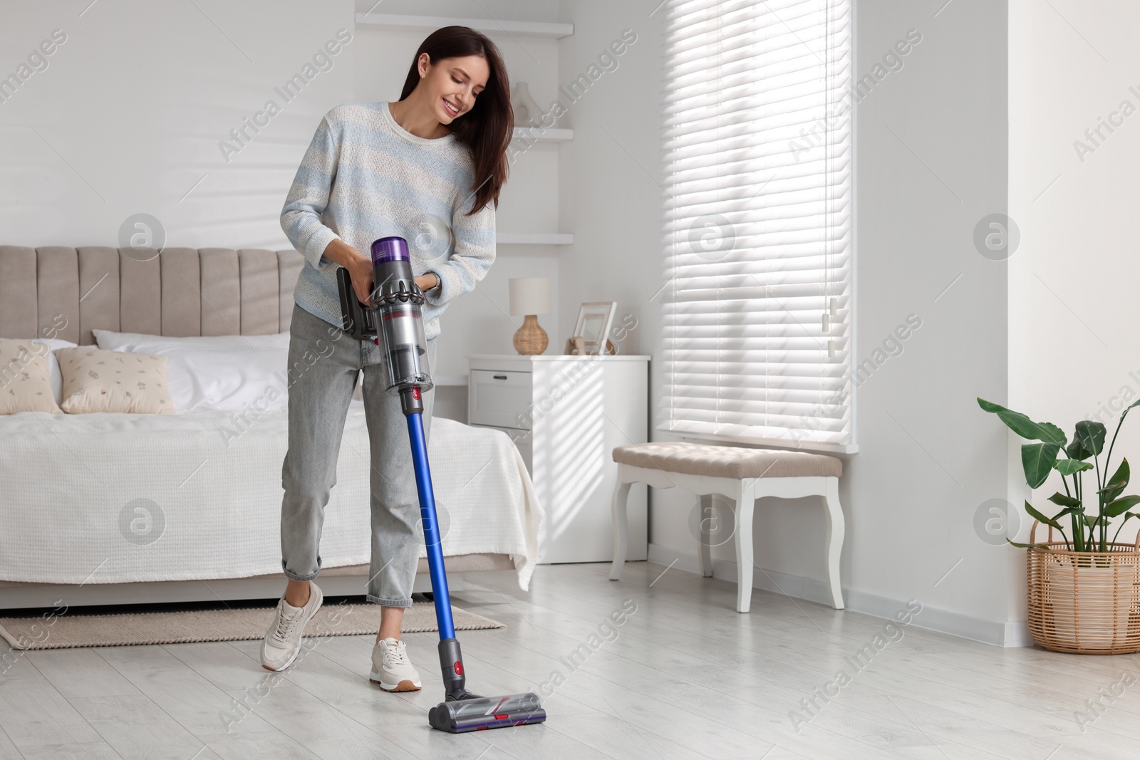 Photo of Smiling young woman cleaning floor with cordless vacuum cleaner in bedroom