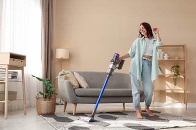 Smiling young woman cleaning rug with cordless vacuum cleaner in living room