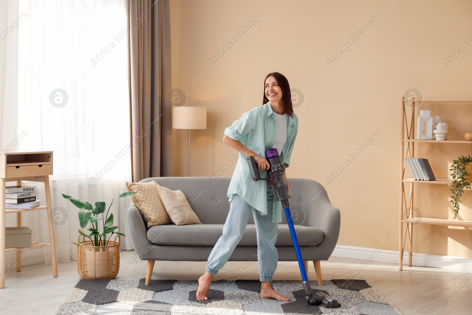 Photo of Smiling young woman cleaning rug with cordless vacuum cleaner in living room