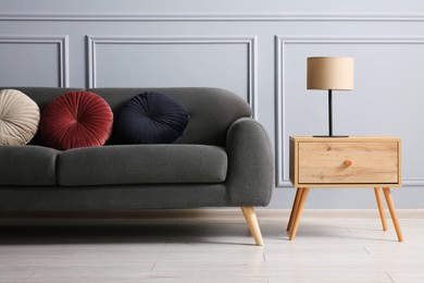 Stylish sofa with cushions and lamp on side table near grey wall