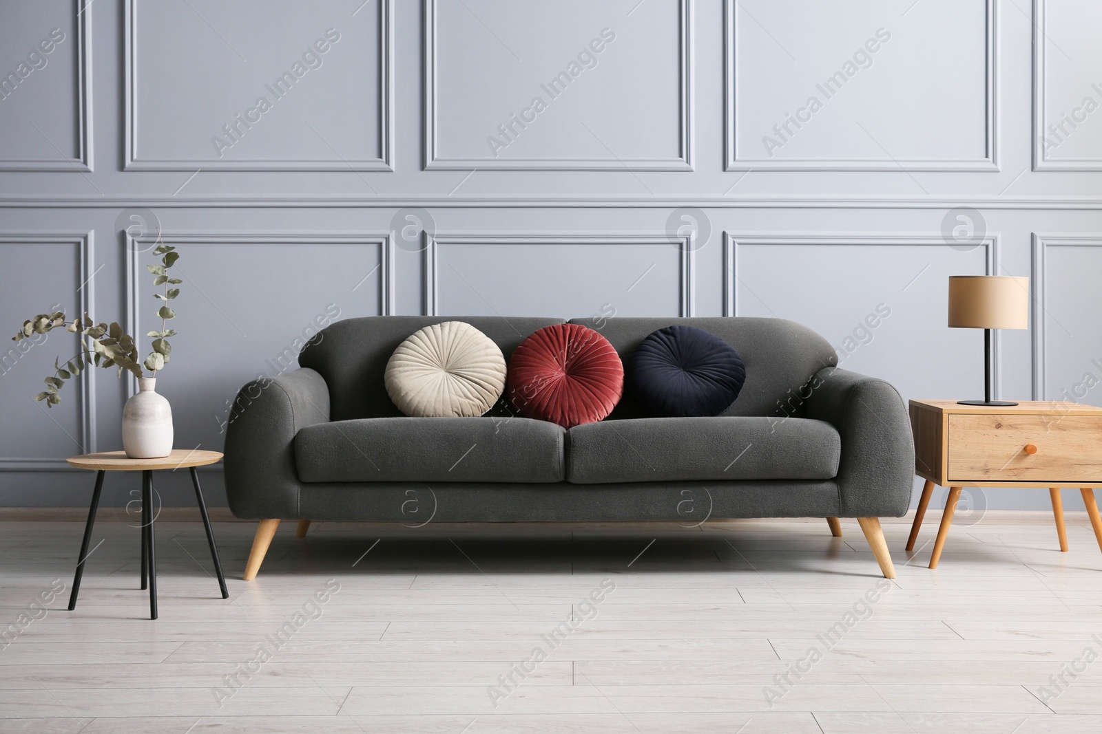 Photo of Stylish sofa with cushions, lamp and eucalyptus branches in vase on side tables in room