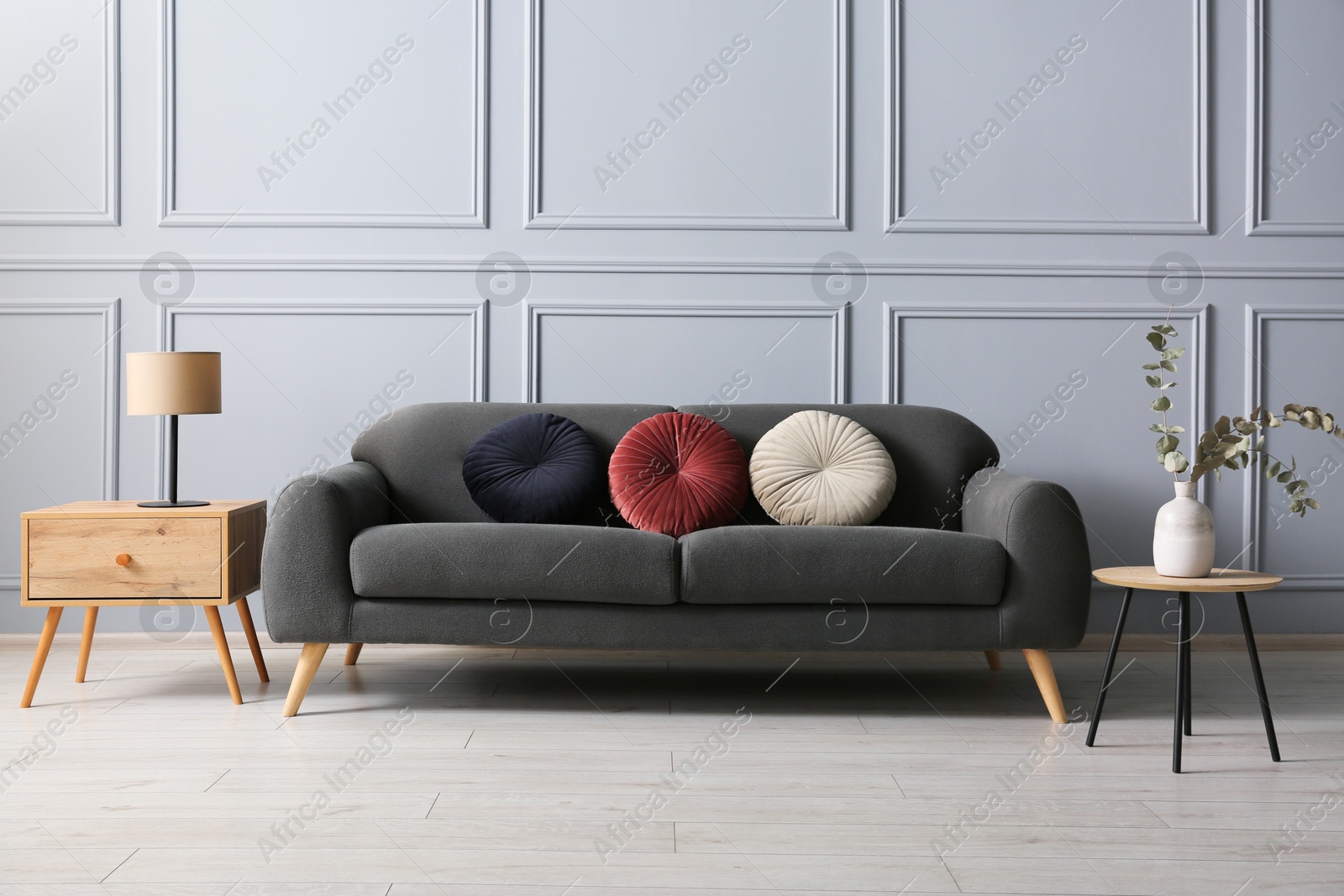 Photo of Stylish sofa with cushions, lamp and eucalyptus branches in vase on side tables in room