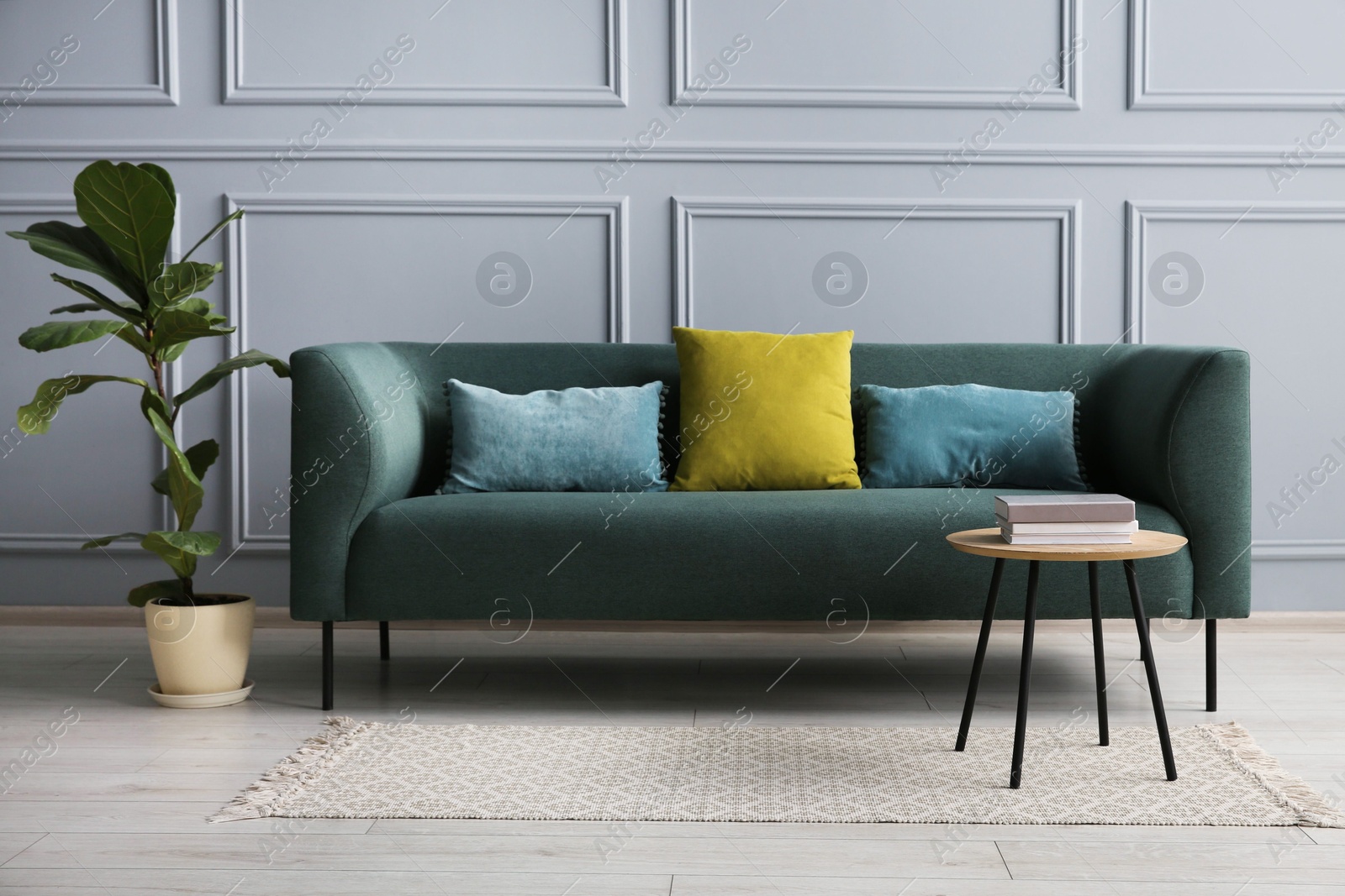 Photo of Stylish sofa with cushions, green plant and books on side table near grey wall