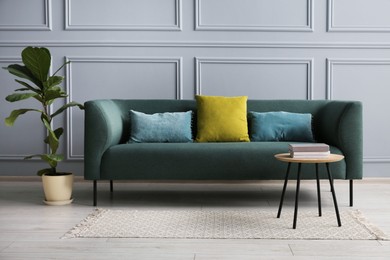 Photo of Stylish sofa with cushions, green plant and books on side table near grey wall