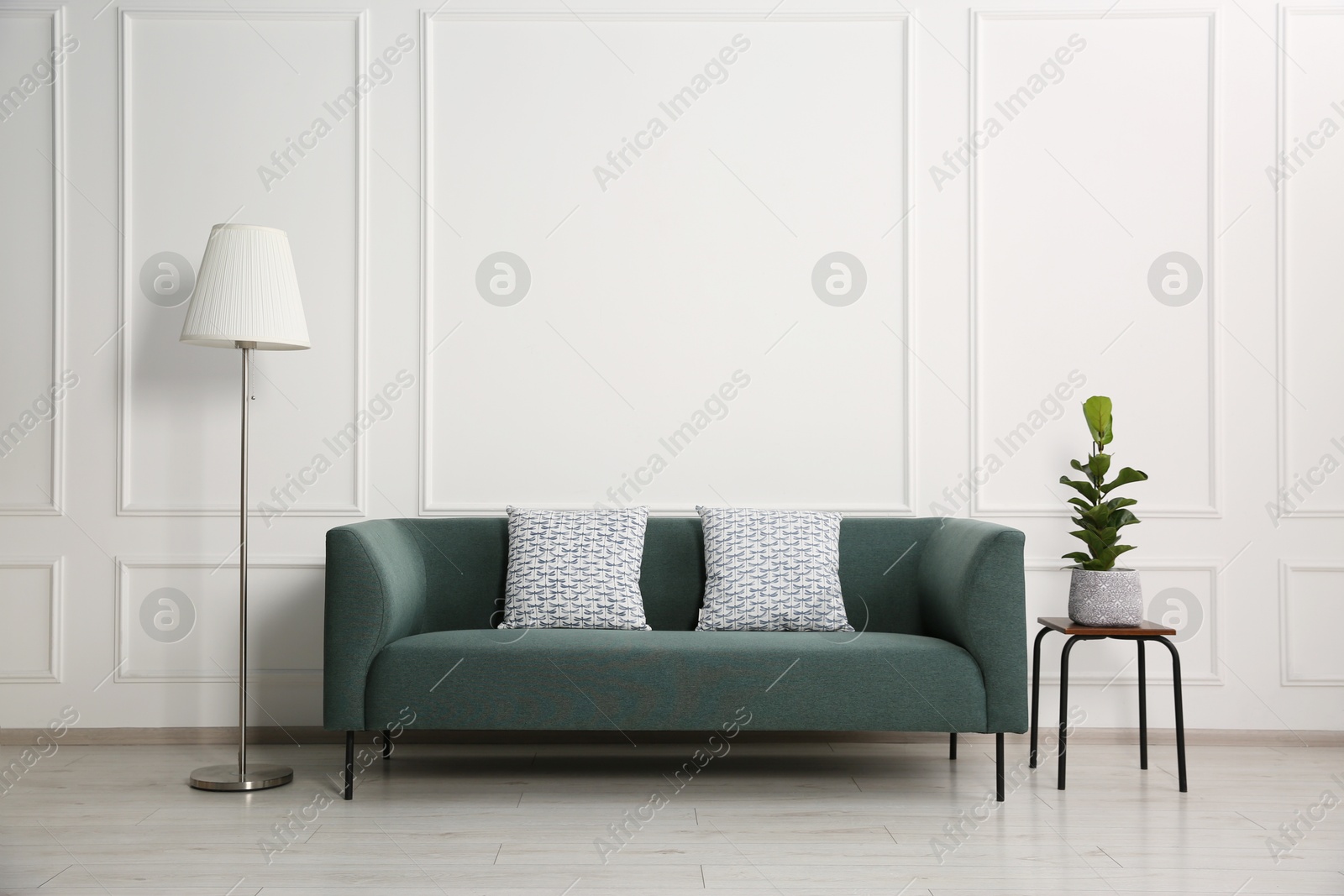 Photo of Stylish sofa with cushions, floor lamp and plant on side table near white wall