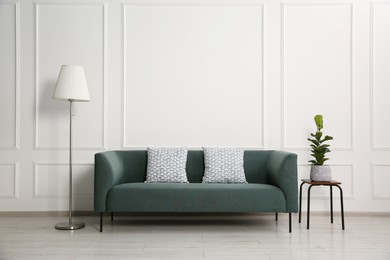 Photo of Stylish sofa with cushions, floor lamp and plant on side table near white wall