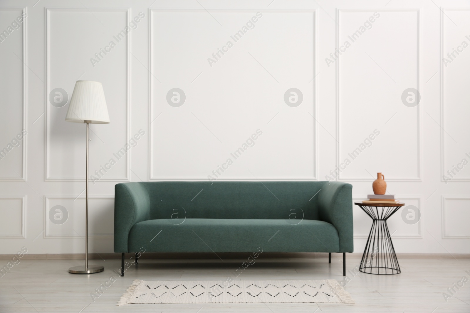 Photo of Stylish sofa, floor lamp and books on side table near white wall