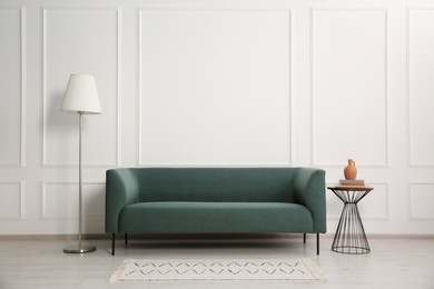 Photo of Stylish sofa, floor lamp and books on side table near white wall