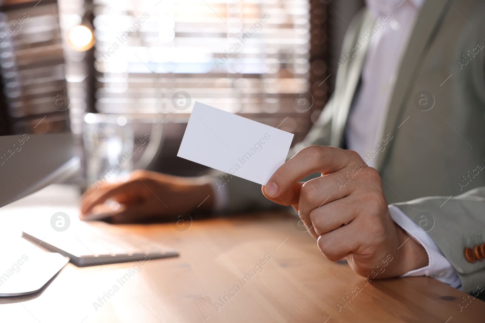 Photo of Man holding blank business card while working with computer at table in office, closeup. Mockup for design