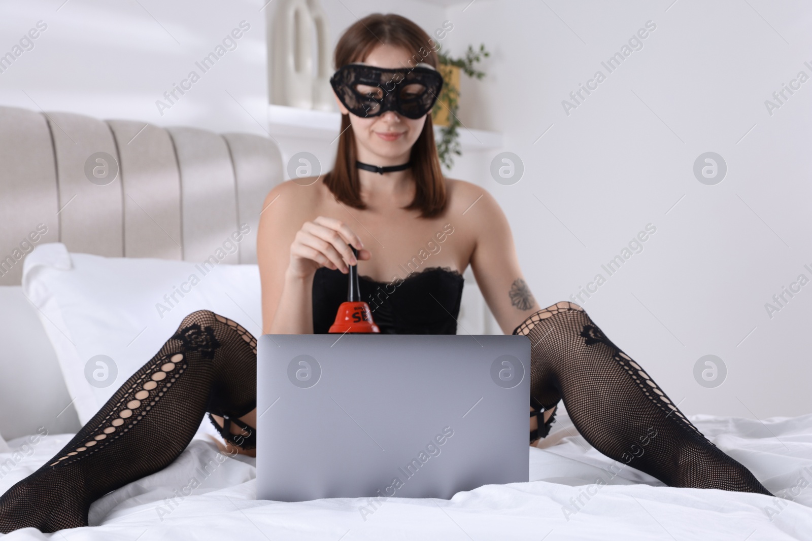 Photo of Prostitution. Webcam model in mask holding bell in front of laptop on bed indoors