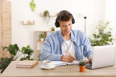 Photo of Student in headphones taking notes while studying with laptop at wooden table indoors