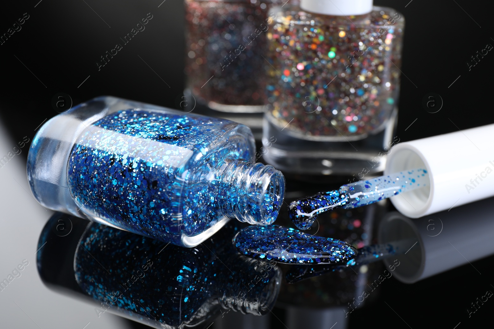 Photo of Glitter nail polishes in bottles and brush on black mirror surface, closeup