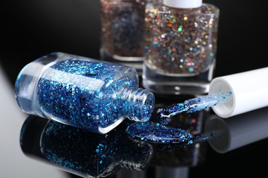 Photo of Glitter nail polishes in bottles and brush on black mirror surface, closeup