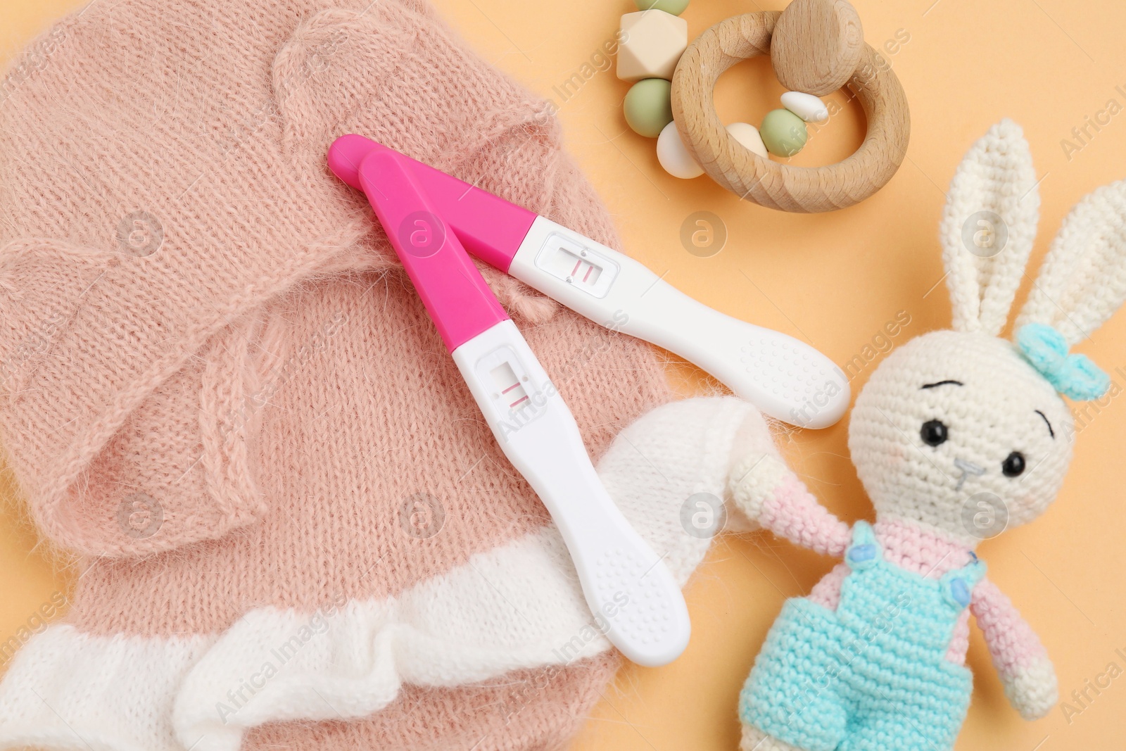 Photo of Pregnancy tests, rattle, toy bunny and baby clothes on pale orange background, above view