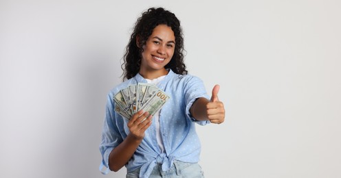 Happy woman with dollar banknotes showing thumbs up on light grey background