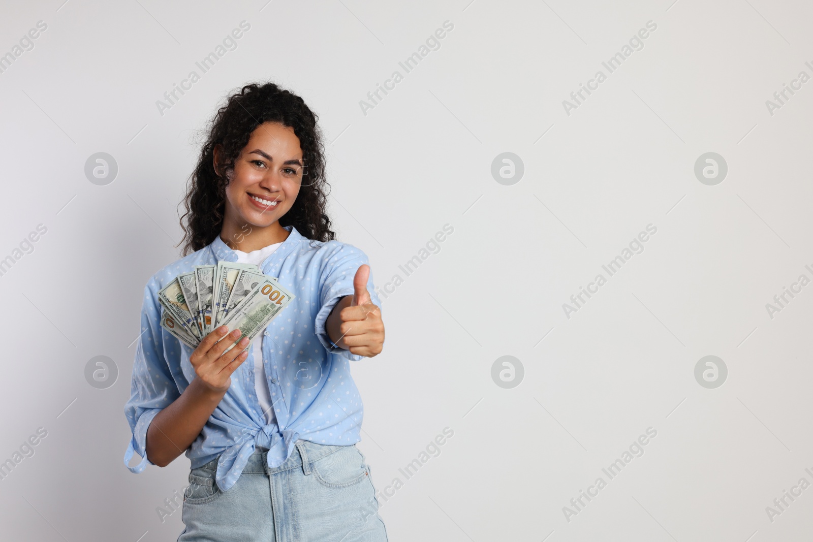 Photo of Happy woman with dollar banknotes showing thumbs up on light grey background, space for text