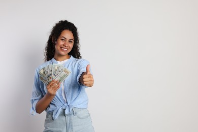 Happy woman with dollar banknotes showing thumbs up on light grey background, space for text