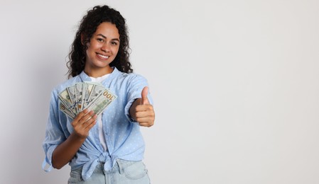 Happy woman with dollar banknotes showing thumbs up on light grey background, space for text