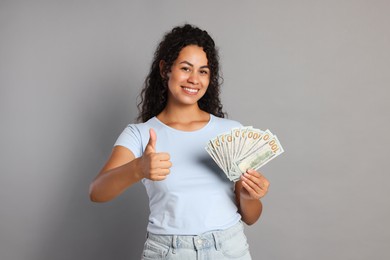 Happy woman with dollar banknotes showing thumbs up on grey background