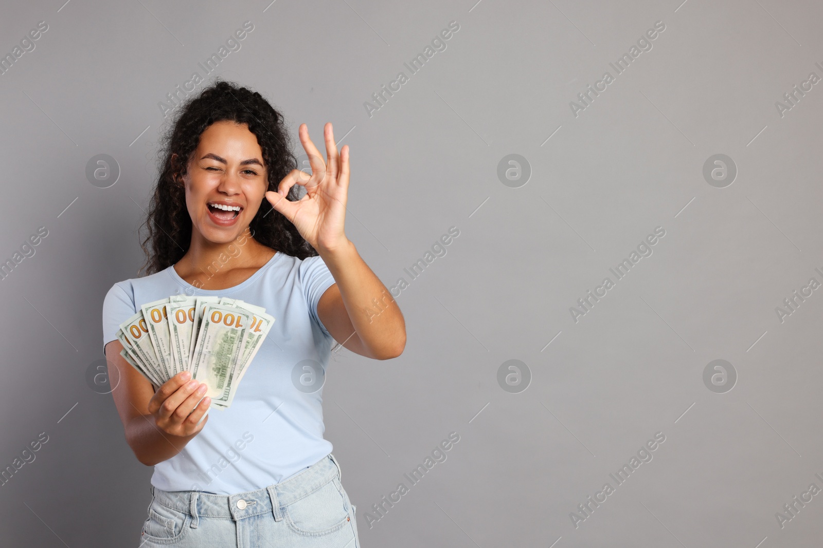 Photo of Happy woman with dollar banknotes showing ok gesture on grey background, space for text