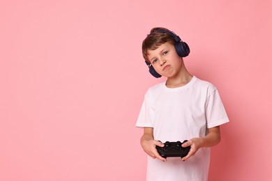 Photo of Cute little boy in headphones playing video game with controller on pink background. Space for text