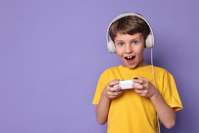 Photo of Happy little boy in headphones playing video game with controller on purple background. Space for text